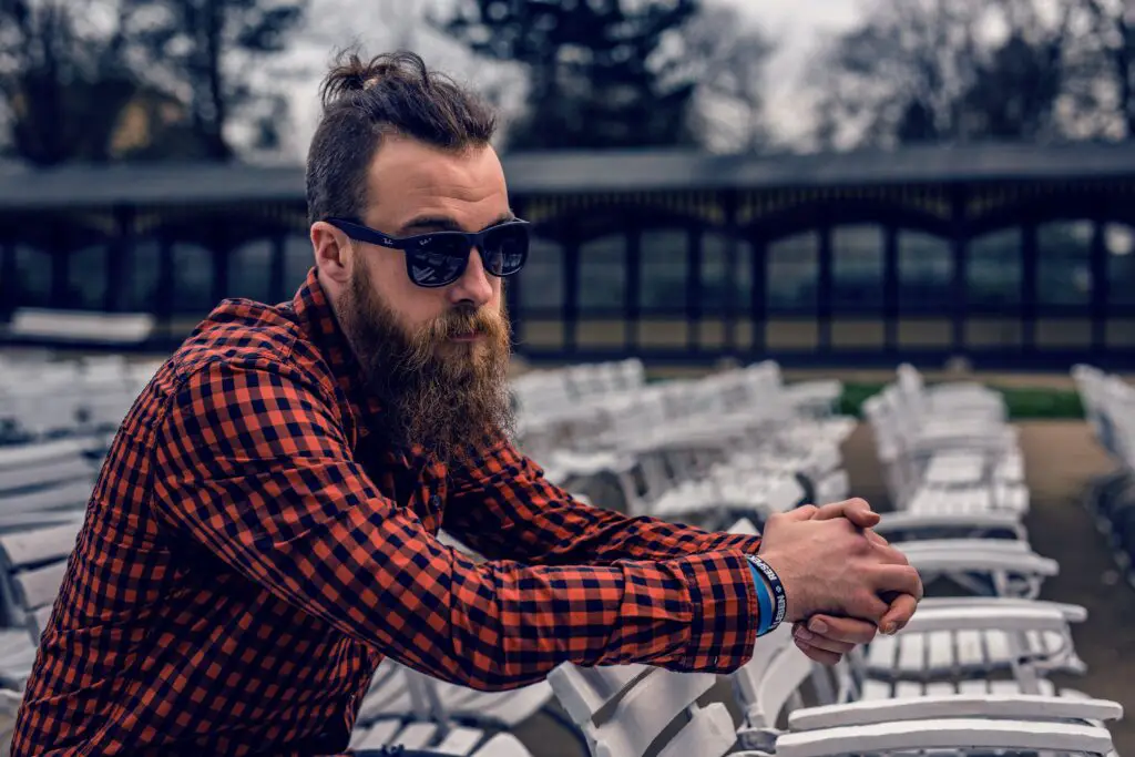 Beard Facts and Myths Hipster man with man bun in red flannel with sunglasses, leaning on a white chair.